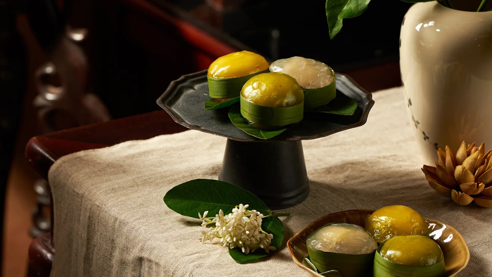 Đĩa bánh trên chân đế đen, bên khăn vải và bình gốm